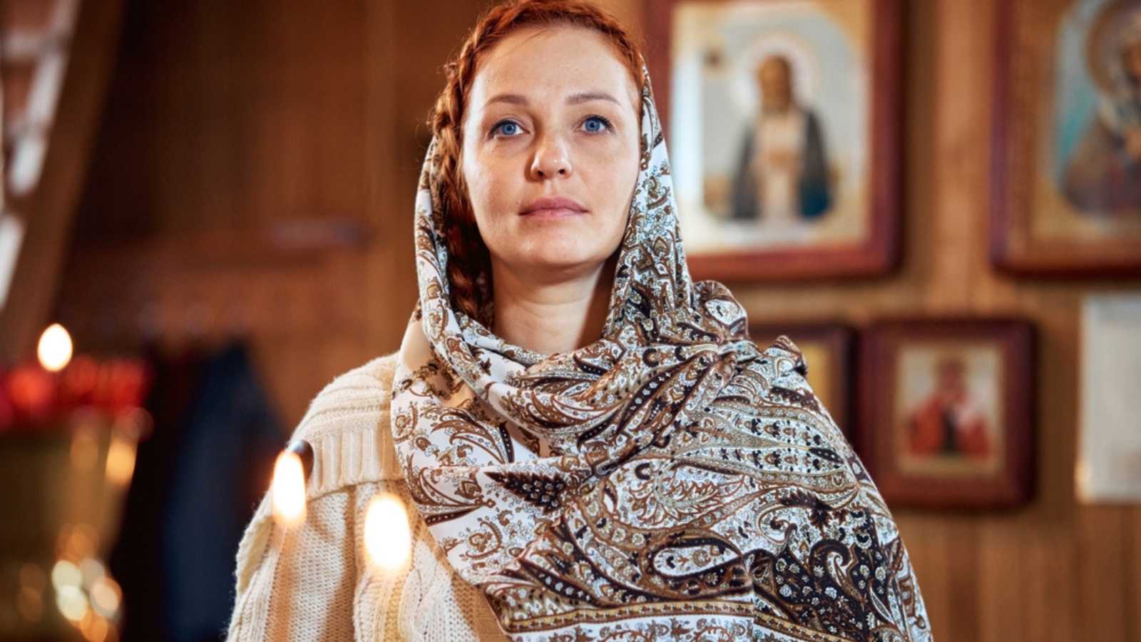 a young woman in a headscarf prays in an Orthodox church and puts candles in front of icons.