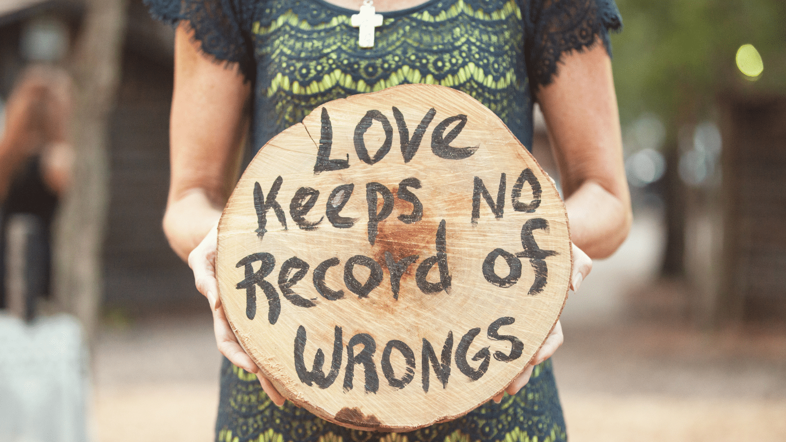 A woman wearing a green dress with a cross necklace, holding a round piece of wood that says, "love keeps no record of wrongs."