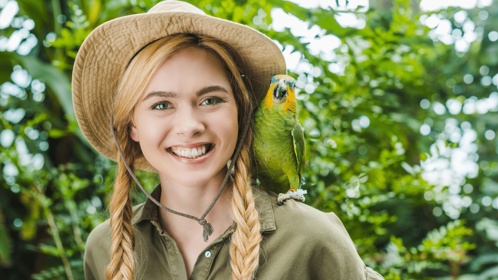 A women with pigtail braids, and a big smile on her face, wearing a green shirt and safari hat, in the jungle with a green and yellow parrot on her shoulder.
