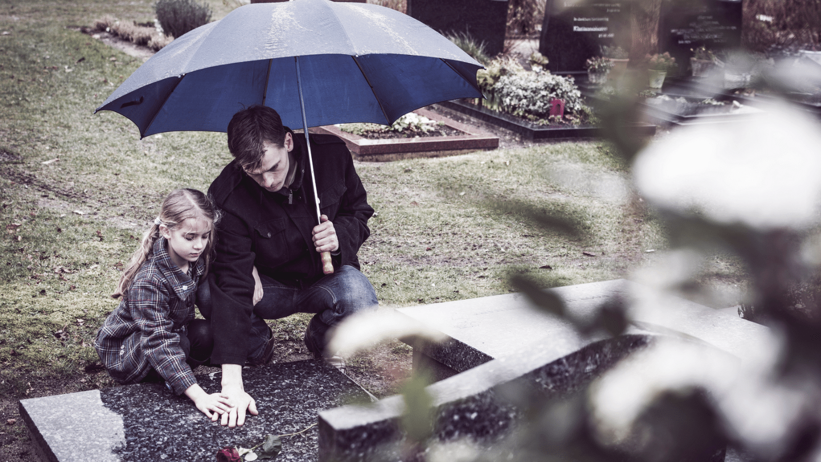 A young father and daughter, at a graveside, holding a blue umbrella, touching a tombstone, mourning a loss.