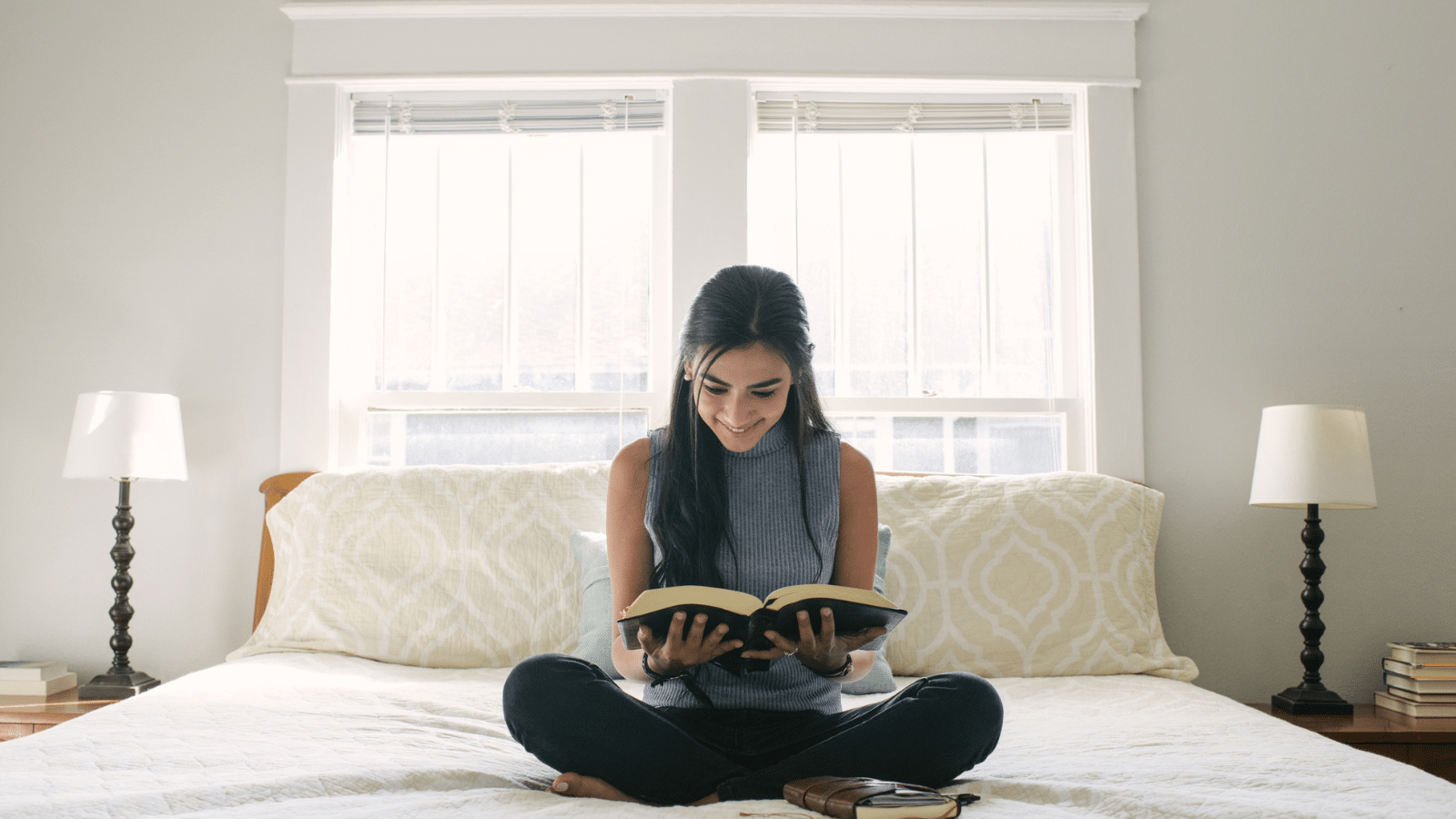 A young woman, sitting on her bed in an all white room, reading her Bible, smiling.