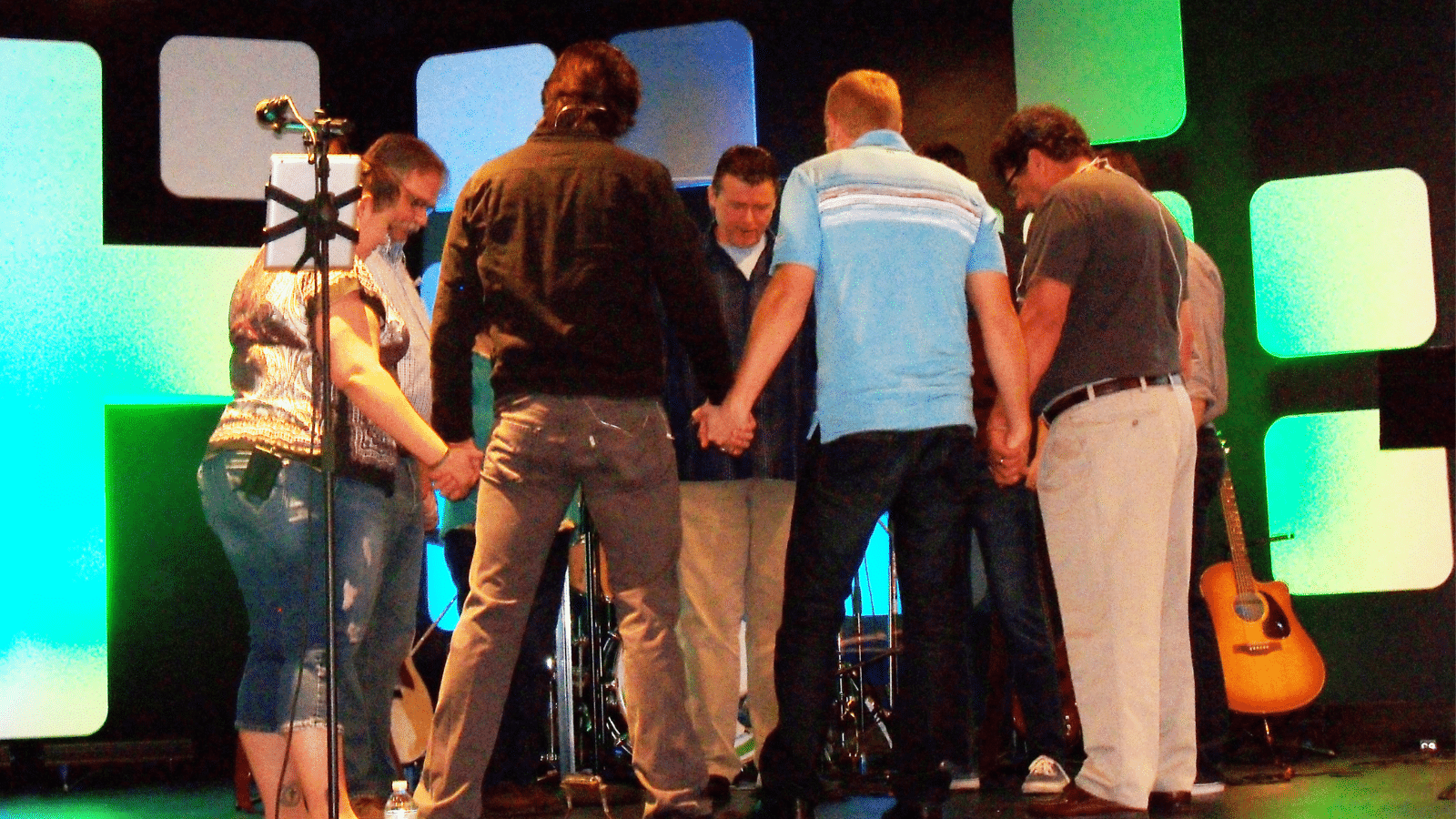 A group of Men and Women gathered together, holding hands in a circle, praying.