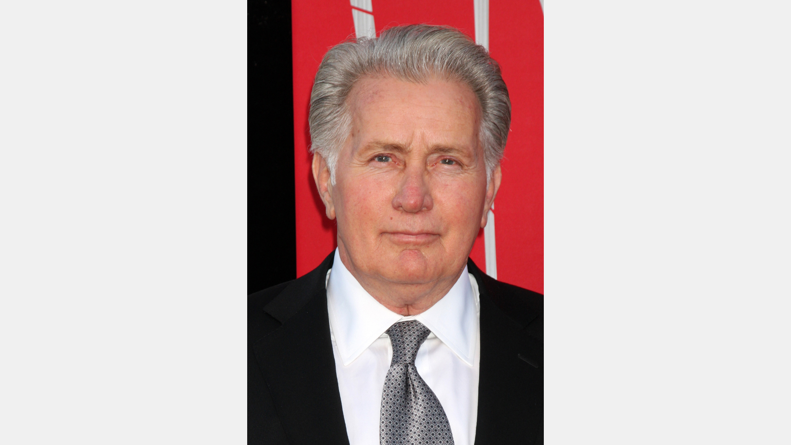 Martin Sheen in a suit in front of a red background.