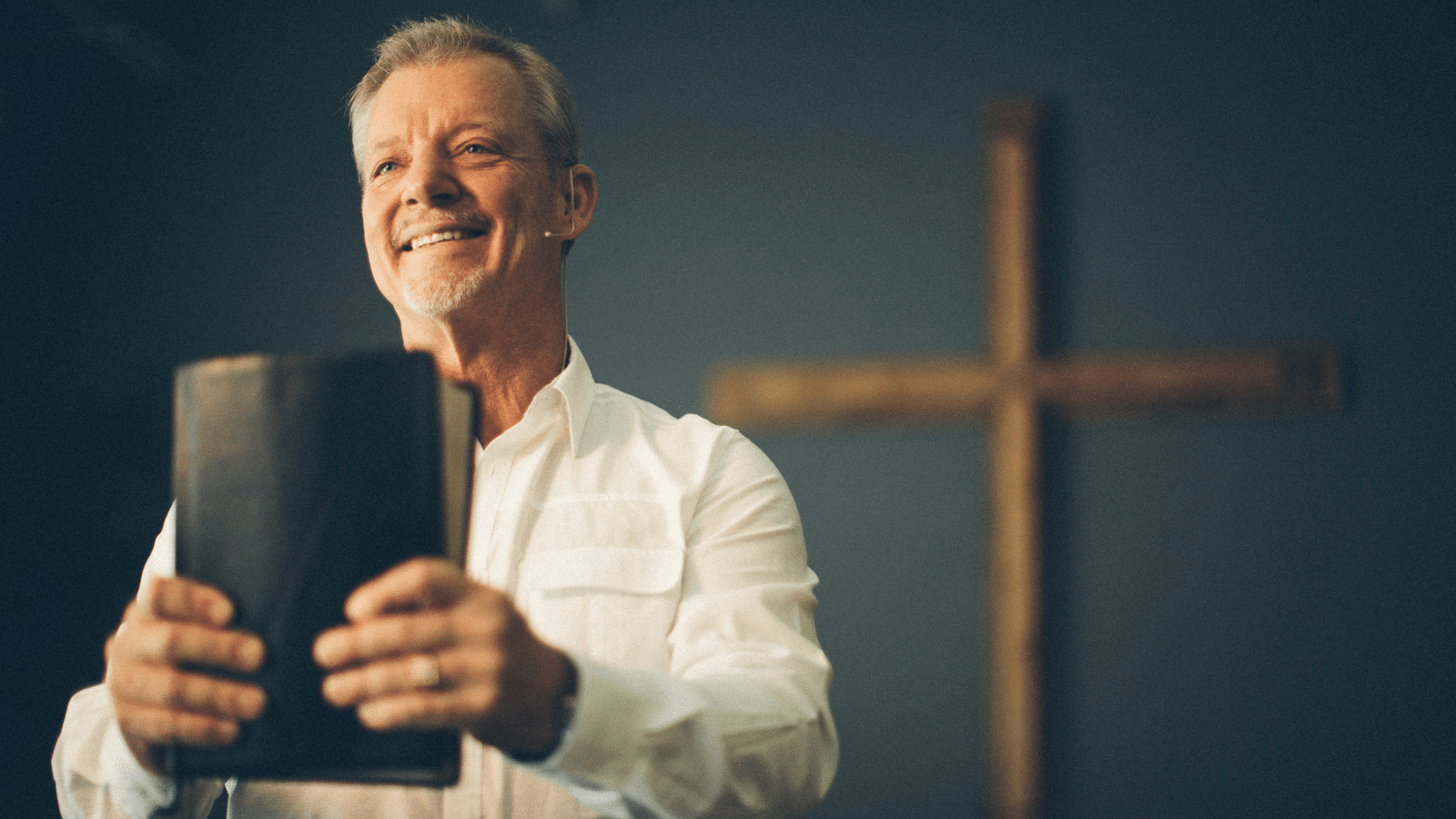 A pastor in front of a cross holding a bible.