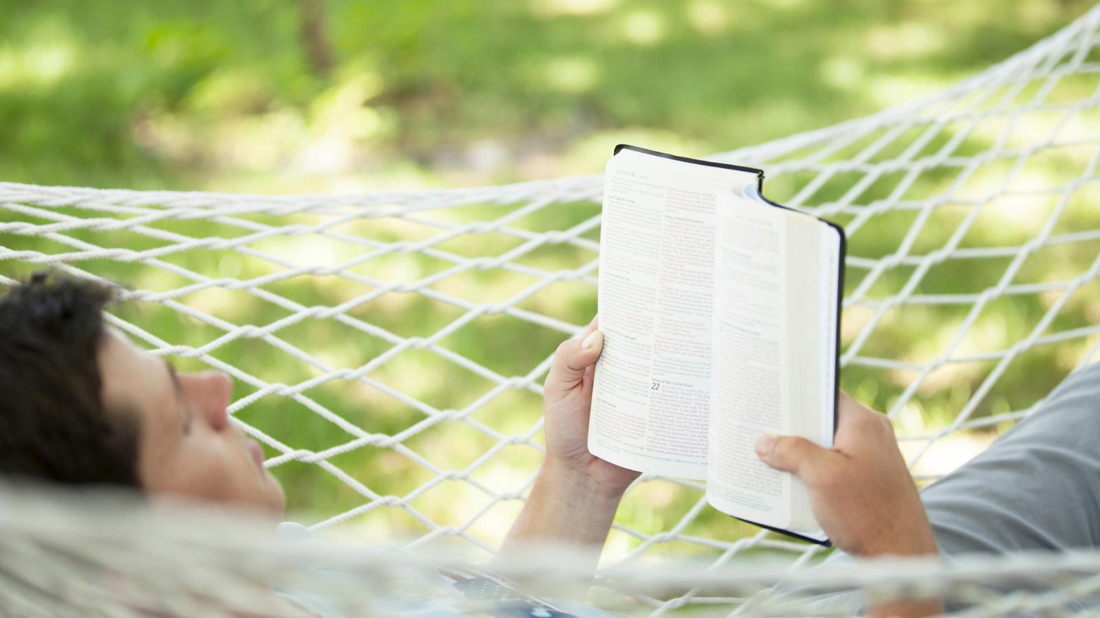A man reading a book while laying in a hammock.