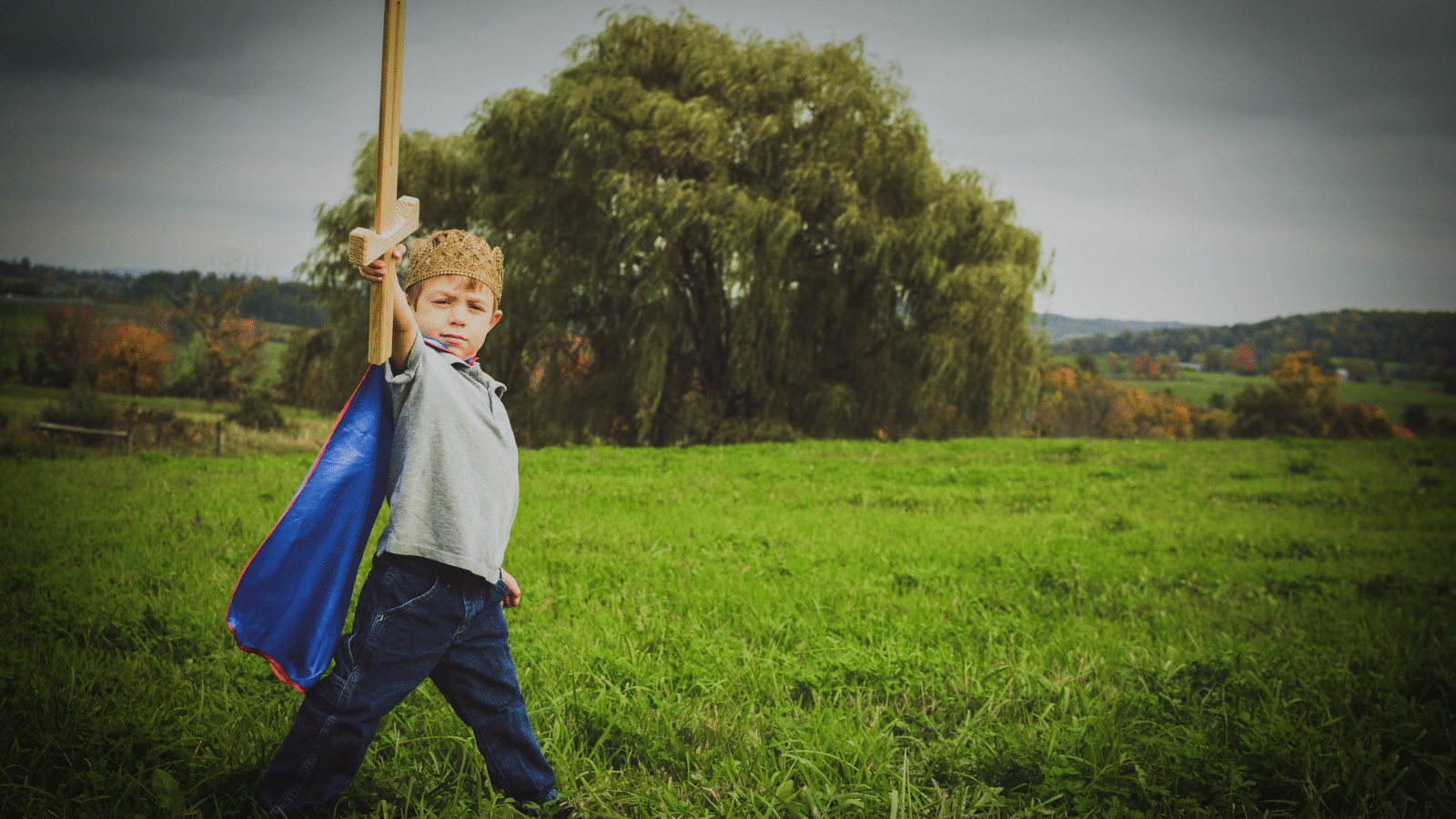 A boy playing outside with a sword and a cape.